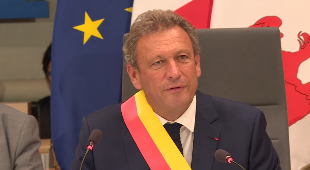 Élu portant une écharpe rouge et jaune, en séance devant les drapeaux européen et régional