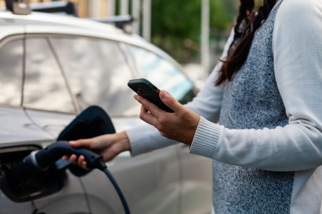 Une personne branche le câble de recharge d'une voiture électrique tout en tenant son smartphone, photo d'illustration